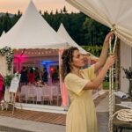 Checkliste Hochzeit wetterfest planen: Eine Frau im gelben Kleid öffnet oder befestigt die Plane eines eleganten, weißen Hochzeitszeltes im Freien.