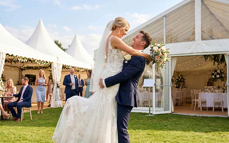 Hochzeit wetterfest planen Ideen: Ehemann hebt Ehefrau glücklich vor dem Hochzeitszelt in die Luft.
