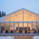 Checkliste Außengastronomie im Winter – Frontalansicht eines modernen, spitz zulaufenden Winterzeltgartens mit weißer Plane und transparenter Glasfront als Erweiterung für ein Restaurant im Schnee.